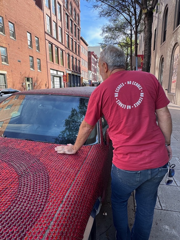Frank, back turned, wearing a brick red NO ECHOES shirt with the circular mark visible between his shoulders, on a sidewalk in downtown Louisville, sunglasses hanging from his back pocket.